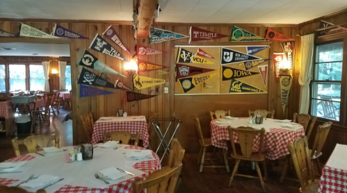 A cozy restaurant interior with wooden walls, checkered tablecloths, and a display of colorful college pennants.