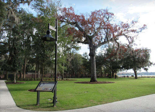 A park scene featuring a large tree with Spanish moss, a historical sign, and a lamp post on a grassy area.