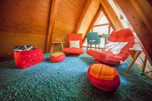 Cozy attic space with wooden walls, colorful chairs, cushions, and a soft blue rug. Bright and inviting atmosphere.