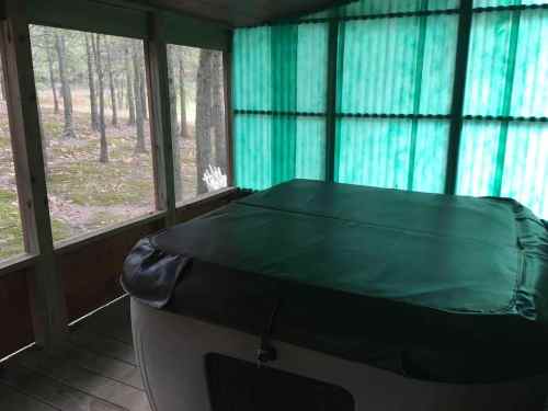 A hot tub with a cover sits in a screened porch surrounded by green-tinted walls and a view of trees outside.