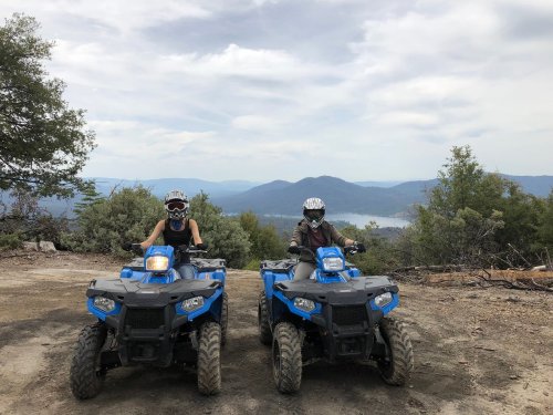 Two people on blue ATVs pose on a dirt path with a scenic mountain view in the background.