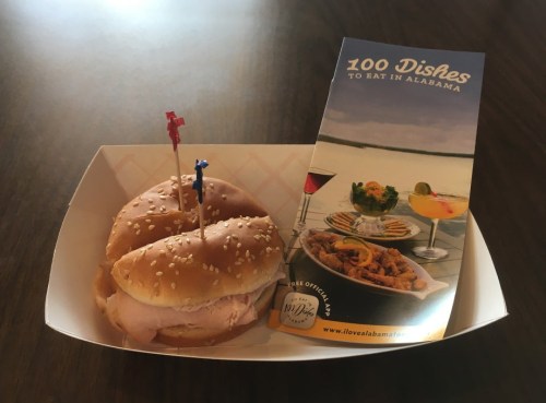 A sesame seed bun sandwich with ham and two toothpicks, next to a brochure titled "100 Dishes to Eat in Alabama."