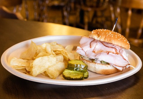 A sesame seed bun sandwich filled with turkey, lettuce, and pickles, served with a side of potato chips.