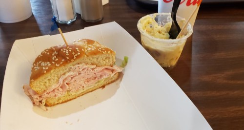 A sesame seed bun sandwich filled with sliced ham, served with a small cup of dessert.