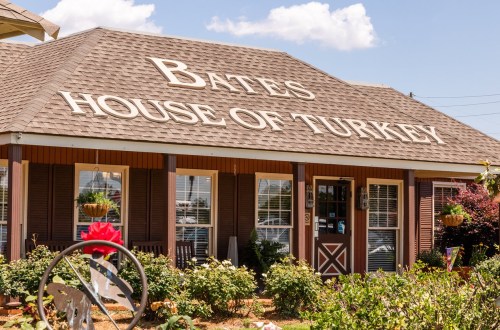 A building with a sign reading "Bates House of Turkey," surrounded by greenery and flowers under a blue sky.