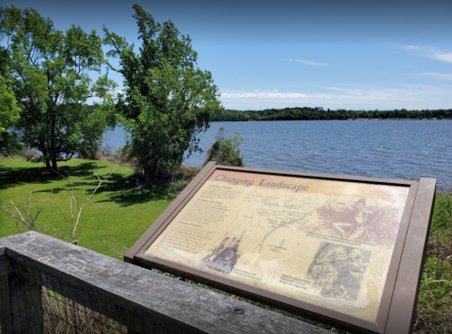 A scenic view of a river with a sign about the changing landscape, surrounded by green trees and grass.