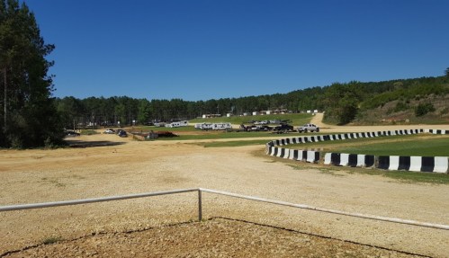 A dirt racetrack with a black and white barrier, surrounded by trees and parked vehicles in the background under a clear blue sky.