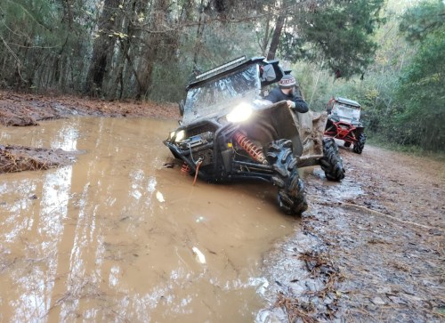 Two off-road vehicles navigating a muddy trail in a forested area, with one partially submerged in water.