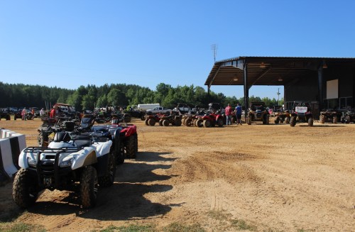 A lineup of ATVs parked on a dirt lot near a large building, with trees and vehicles in the background under a clear blue sky.