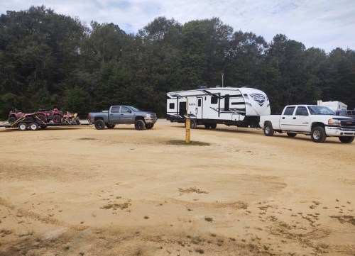 A gravel lot with two trucks and a large RV, surrounded by trees under a cloudy sky.