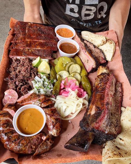 A platter of assorted BBQ meats, including ribs, brisket, sausage, pickles, onions, and sauces, served with tortillas.