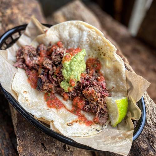 A close-up of a taco filled with shredded meat, salsa, guacamole, and a lime wedge, served on a paper-lined basket.