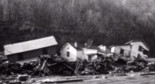 Black and white image of collapsed houses and debris in a rural area, surrounded by trees.