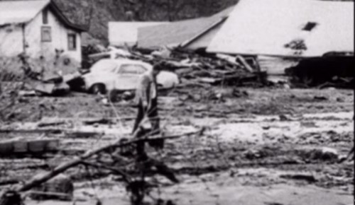 A person stands in a muddy area near damaged houses and a submerged car after a natural disaster.