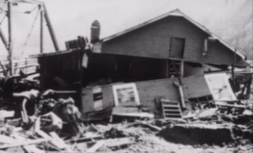 A damaged house tilted on its foundation, surrounded by debris and destruction from a natural disaster.
