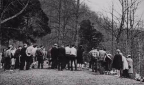 A group of people gathered outdoors in a wooded area, facing away from the camera, with trees in the background.