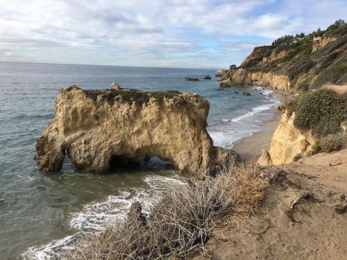 A rocky coastline with a natural arch, sandy beach, and gentle waves under a cloudy sky.
