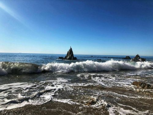 A serene beach scene with gentle waves crashing against rocky formations under a clear blue sky.