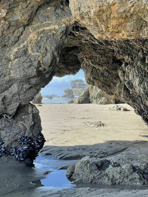A rocky arch frames a sandy beach with gentle waves and distant cliffs under a clear sky.