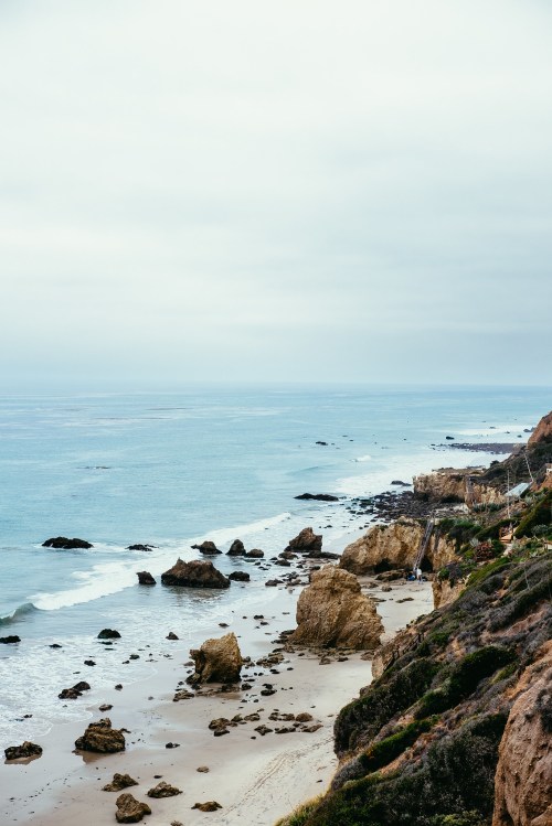 A serene coastal view featuring rocky cliffs, sandy beach, and calm ocean waters under a cloudy sky.