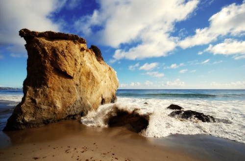 A rocky cliff stands by the shore, with waves crashing against it under a blue sky with scattered clouds.