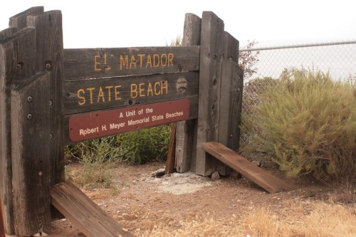 Sign for El Matador State Beach, featuring wooden posts and a sandy area with shrubs in the background.