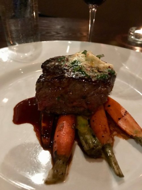 A beautifully plated steak topped with herb butter, served on a bed of roasted carrots and drizzled with sauce.