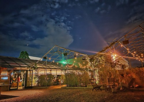 A cozy greenhouse at night, illuminated by string lights, with people relaxing outside under a cloudy sky.