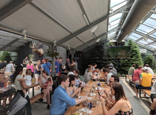 A bustling indoor dining space with people enjoying meals at long tables, surrounded by greenery and a mounted deer head.