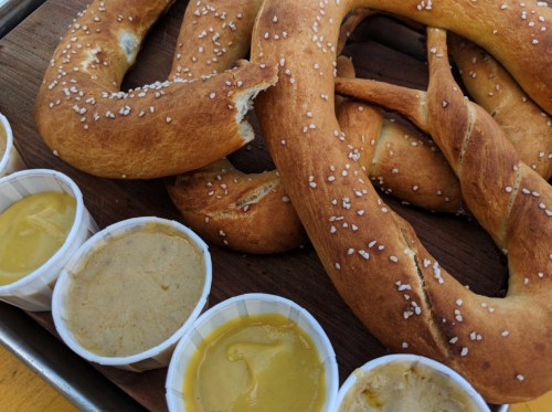 A close-up of soft pretzels with coarse salt, served with small cups of mustard and cheese dip on a wooden platter.
