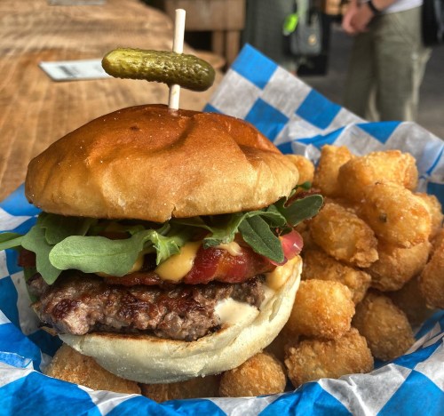 A juicy burger with bacon, cheese, and greens, served with crispy tater tots and a pickle on top.