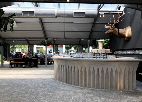 A modern bar with a curved concrete counter, a mounted deer head, and patrons seated in a spacious, open area.