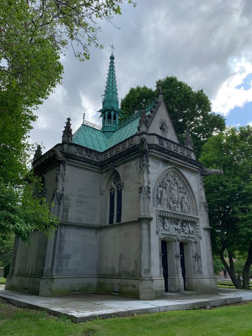 A stone mausoleum with a green spire, intricate carvings, and surrounded by trees under a cloudy sky.