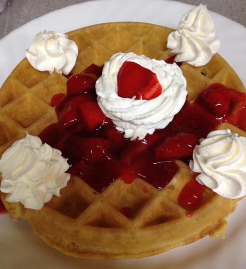 A golden waffle topped with whipped cream and strawberry sauce, garnished with fresh strawberries and dollops of whipped cream.