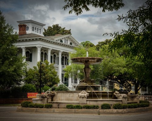 Historic white mansion with columns behind a decorative fountain surrounded by greenery under a cloudy sky.