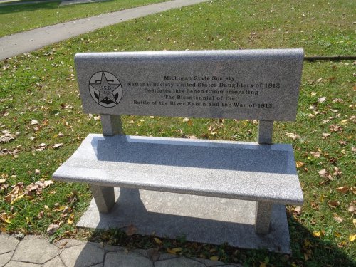 Gray stone bench with a plaque commemorating the Battle of the River Raisin and the War of 1812, surrounded by grass.
