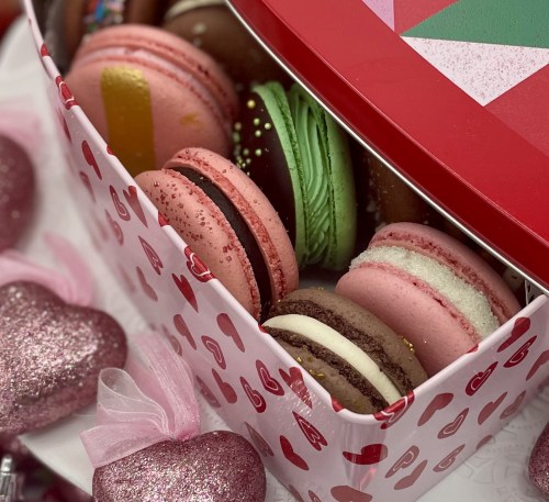 A colorful assortment of macarons in a heart-themed box, surrounded by glittery heart decorations.