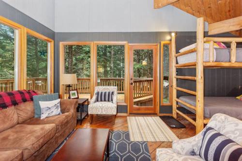 Cozy living room with a sofa, armchair, and bunk beds, featuring large windows overlooking a forested area.