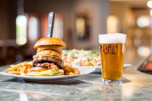 A large burger with onion rings, lettuce, and tomato, served with fries and a pint of beer on a table.