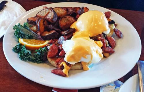 A plate of eggs benedict with sausage, cheese sauce, and seasoned potatoes, garnished with lettuce and an orange slice.