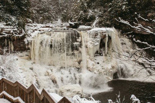 A frozen waterfall surrounded by snow-covered trees and a wooden railing in winter.