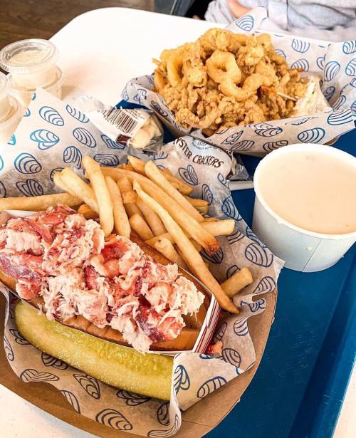 A lobster roll with fries and a pickle, alongside a bowl of clam chowder and fried seafood on a blue tray.