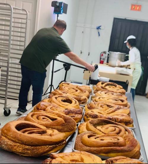 A baker prepares large cinnamon rolls on trays, while another person sets up equipment in a kitchen.