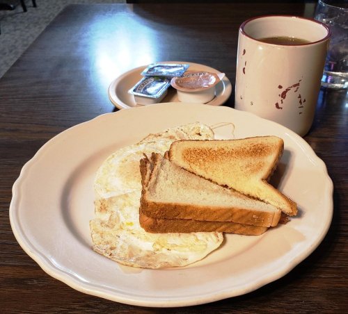 A plate with two fried eggs and two slices of toast, accompanied by a cup of coffee and small condiment packets.