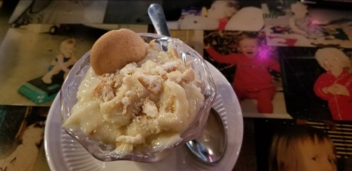 A dessert in a glass bowl topped with crushed cookies and a whole cookie, placed on a table with childhood photos.