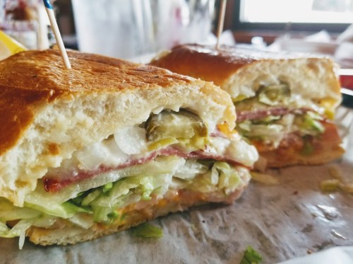 Close-up of a sandwich with layers of lettuce, pickles, and meats, served on a toasted bun with toothpicks.