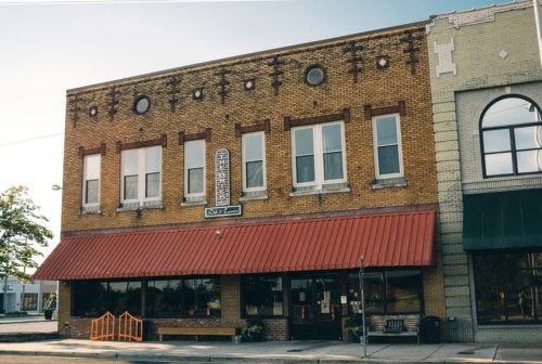 A brick building with a red awning, featuring large windows and decorative elements, in a small town setting.