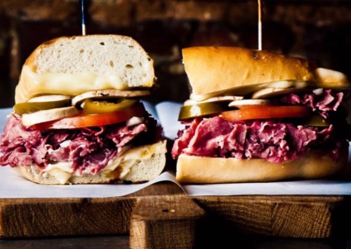 A close-up of a sandwich with layers of roast beef, cheese, pickles, tomatoes, and onions on a soft bun.