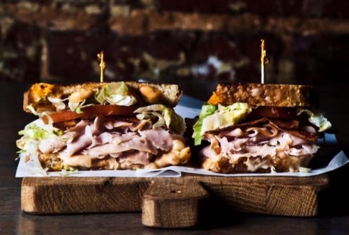 A close-up of a stacked sandwich with layers of turkey, ham, lettuce, and tomato, served on a wooden board.