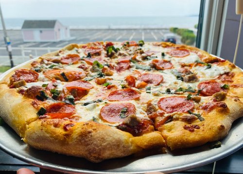 A freshly baked pepperoni pizza topped with herbs, sitting on a silver tray with a scenic view of the ocean in the background.